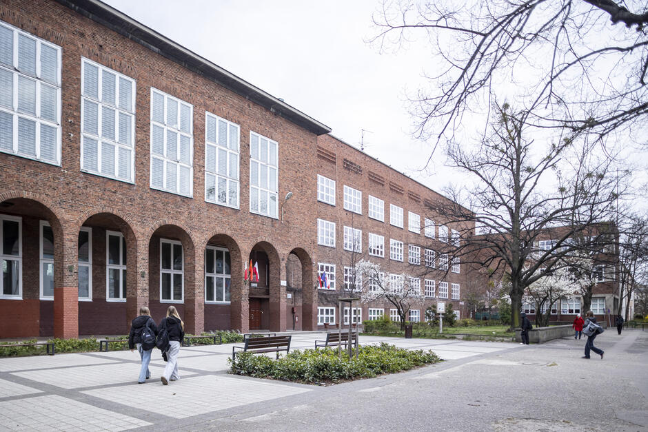 Na zdjęciu widoczny jest duży, kilkupiętrowy budynek wykonany z czerwonej cegły, który wygląda na szkołę lub inną instytucję edukacyjną. Charakterystycznym elementem architektury są wysokie, łukowe okna na parterze oraz liczne, białe ramy okienne na wyższych piętrach. Przy wejściu do budynku powiewają flagi, co może sugerować, że jest to placówka międzynarodowa lub publiczna. Przed budynkiem znajduje się zadbany dziedziniec z wybrukowanymi alejkami, ławkami oraz niewielkimi rabatami roślinnymi. Wokół rosną drzewa, niektóre z nich właśnie kwitną, co wskazuje na wczesną wiosnę. Na zdjęciu widać kilka osób – prawdopodobnie uczniów – wchodzących lub wychodzących z budynku, co potęguje wrażenie, że jest to miejsce nauki.