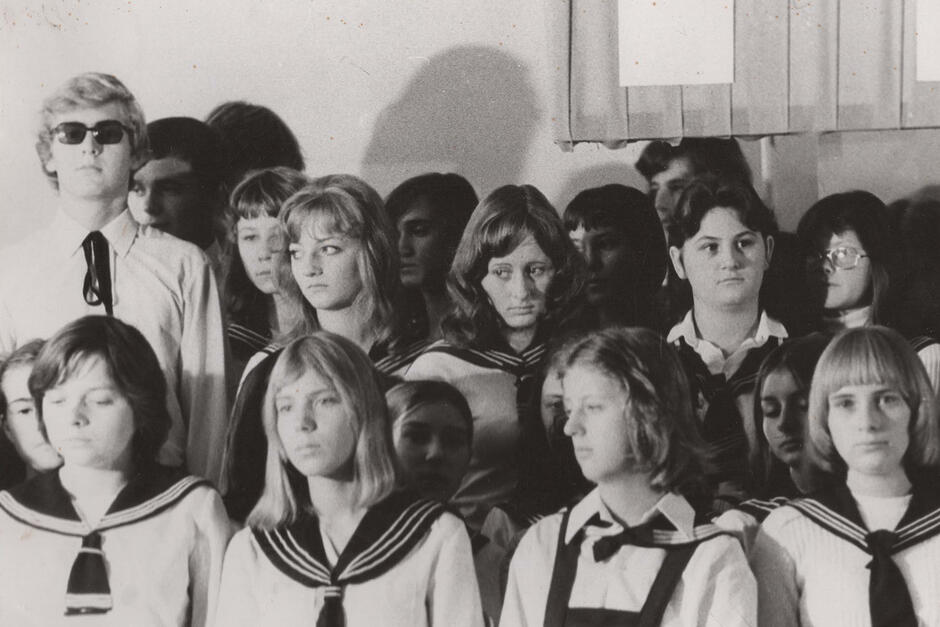 To czarno-białe zdjęcie przedstawia grupę młodych osób, prawdopodobnie uczniów, ustawionych do wspólnej fotografii, być może klasowej lub z okazji szkolnej uroczystości. Uczniowie są ubrani w jednakowe stroje – dziewczęta mają na sobie bluzki z charakterystycznymi kołnierzami marynarskimi i krawatami, co sugeruje, że zdjęcie zostało wykonane w kraju, gdzie taki ubiór był częścią szkolnego mundurka (możliwe, że w Polsce lub innym kraju Europy Środkowo-Wschodniej, w okresie PRL-u). Jeden z chłopców nosi ciemne okulary, co wyróżnia go na tle reszty grupy.