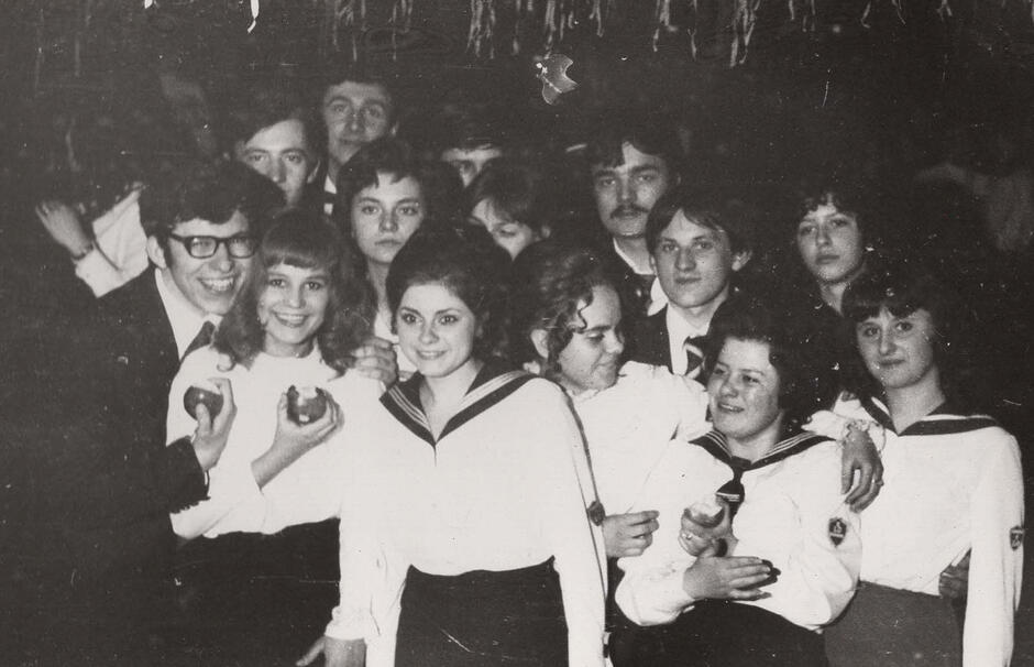 To czarno-białe zdjęcie przedstawia grupę młodzieży, prawdopodobnie uczniów szkoły średniej, a może nawet studentów niższych lat, zgromadzonych na jakimś oficjalnym wydarzeniu – być może rozpoczęciu lub zakończeniu roku szkolnego, akademii szkolnej czy innej uroczystości. Dziewczęta ubrane są w charakterystyczne mundurki z marynarskimi kołnierzami – typowe dla uczennic w Polsce i krajach bloku wschodniego w latach 60., 70. i 80. XX wieku. Chłopiec w centrum ma na sobie garnitur i krawat z dużym wzorem, również bardzo w stylu lat 70. Atmosfera jest raczej luźna – niektórzy się uśmiechają, inni patrzą gdzieś w bok lub rozmawiają, co sugeruje moment oczekiwania lub przerwy między oficjalnymi punktami wydarzenia.