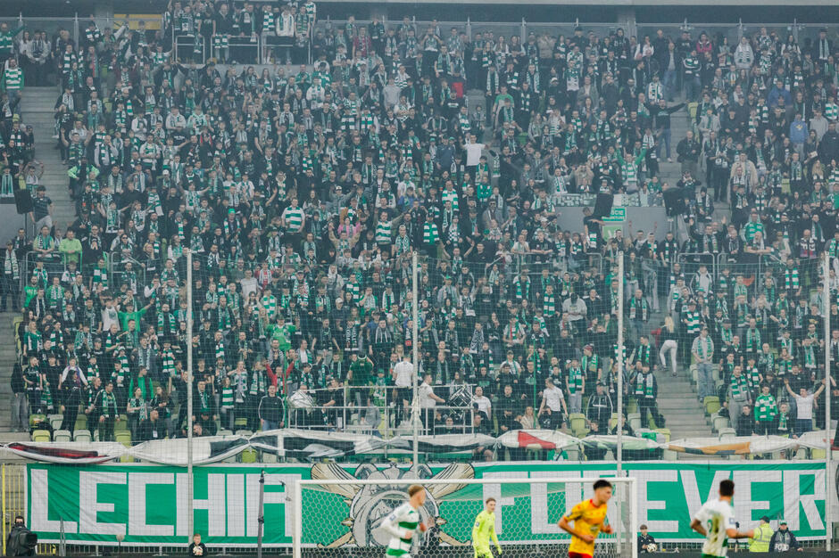 Duży tłum ludzi na stadionie. Ludzie noszą zielone i białe koszulki pasowe i stoją na trybunach. Wszyscy trzymają telefony i wydają się dopingować lub świętować. Na pierwszym planie znajduje się bramka piłkarska z napisem LECHIEVER nad nią. Dwóch piłkarzy z drużyn przeciwnych stoi na boisku przed bramką, jeden w żółtej koszuli, a drugi w pomarańczowej koszuli. Jest też kilku innych piłkarek w żylakach. Stoiska są wypełnione widzami, niektórzy stoją, i niektóre siedzą, oraz banery oraz flagi wiszące z biegunów. Wygląda na to, że obraz został zrobiony podczas meczu piłki nożnej.