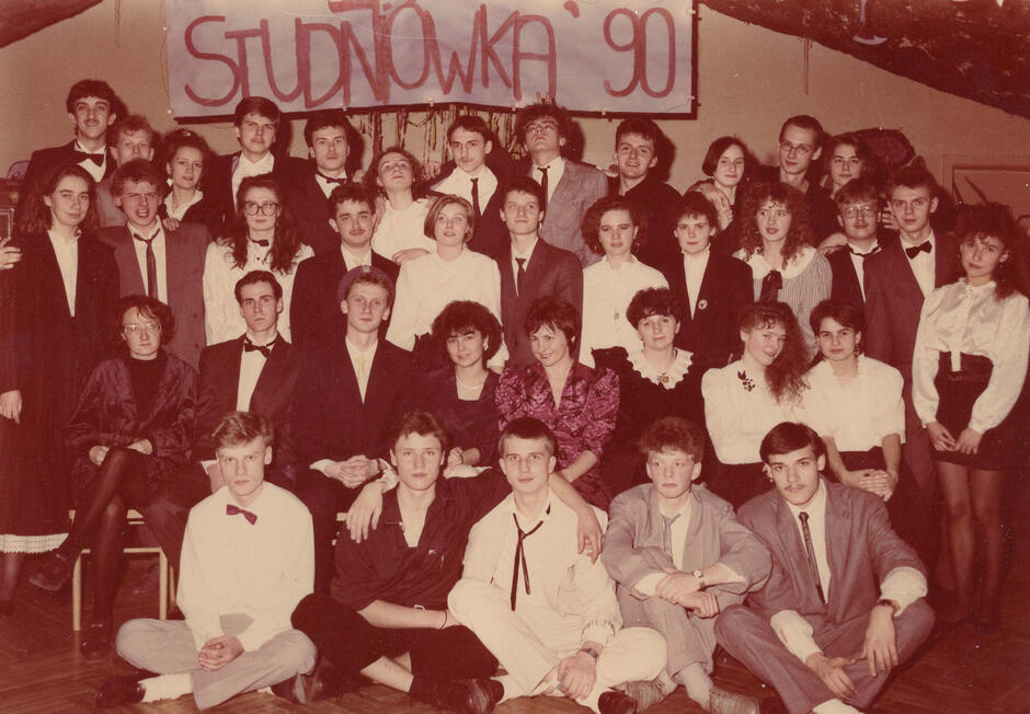 To zdjęcie pochodzi ze studniówki w 1990 roku, co potwierdza duży transparent z napisem „STUDNIÓWKA '90” widoczny w tle. Zdjęcie przedstawia grupę uczniów (zapewne klasy maturalnej) wraz z nauczycielami – prawdopodobnie wychowawcą lub dyrekcją – pozujących do pamiątkowej fotografii podczas balu studniówkowego.