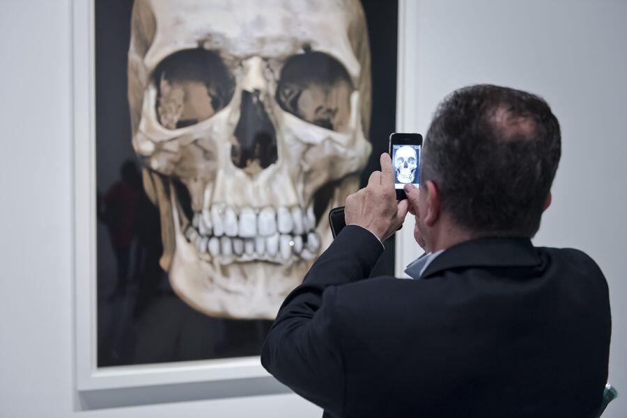 Mężczyzna w garniturze fotografuje dużą grafikę przedstawiającą realistyczny obraz ludzkiej czaszki zawieszoną w galerii sztuki. Kompozycja ukazuje interakcję widza ze sztuką przez pryzmat nowoczesnych technologii, jak telefon komórkowy