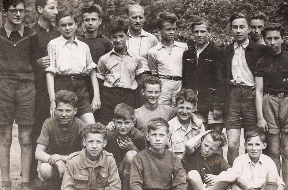 Na tej czarno-białej fotografii widzimy grupę chłopców, prawdopodobnie uczestników obozu harcerskiego, kolonii letnich lub szkolnego wyjazdu, wraz z jednym dorosłym opiekunem (widocznym w środkowej górnej części zdjęcia). Zdjęcie ma typowo letni, rekreacyjny charakter – chłopcy są ubrani swobodnie: w krótkie spodenki, koszule z podwiniętymi rękawami lub t-shirty.