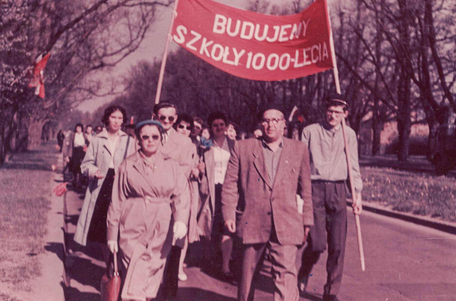 To kolorowe zdjęcie z lat 60. XX wieku przedstawia grupę ludzi uczestniczących w pochodzie lub manifestacji społecznej, związanej z akcją budowy szkół w ramach programu „Tysiąc szkół na Tysiąclecie Państwa Polskiego”.