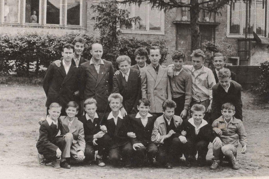 To czarno-białe zdjęcie przedstawia grupę chłopców, prawdopodobnie uczniów starszych klas szkoły podstawowej lub szkoły zawodowej, wraz z nauczycielem lub wychowawcą. Zdjęcie zostało wykonane na zewnątrz – najpewniej na dziedzińcu szkolnym lub placu przyinternatowym.