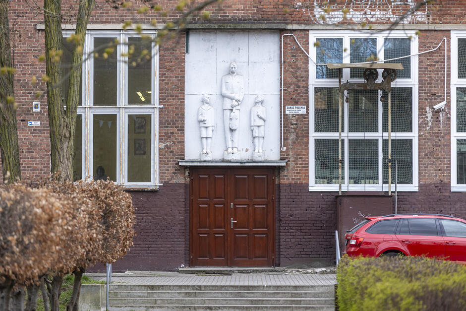 To zdjęcie przedstawia wejście do budynku szkolnego – charakterystycznego dla architektury szkół budowanych w czasach PRL, prawdopodobnie w ramach programu Tysiąclatek, czyli „Szkół Tysiąclecia”.