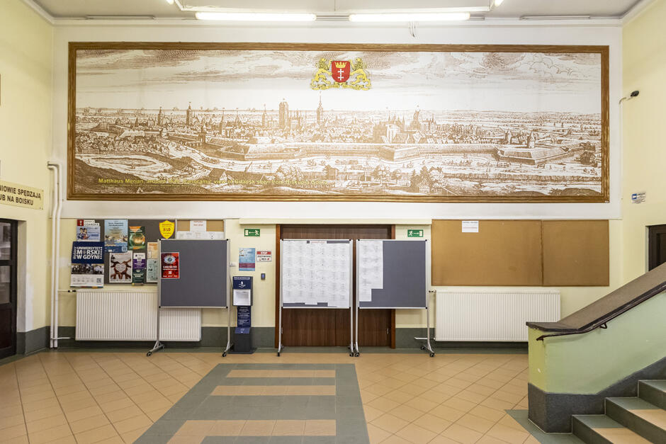 To zdjęcie przedstawia wnętrze szkoły, konkretnie główny hol lub foyer, prawdopodobnie na parterze, przy wejściu głównym. Uwagę przykuwa duża grafika na ścianie, przedstawiająca panoramiczny widok Gdańska autorstwa Matthäusa Meriana – znanego rytownika i kartografa z XVII wieku.