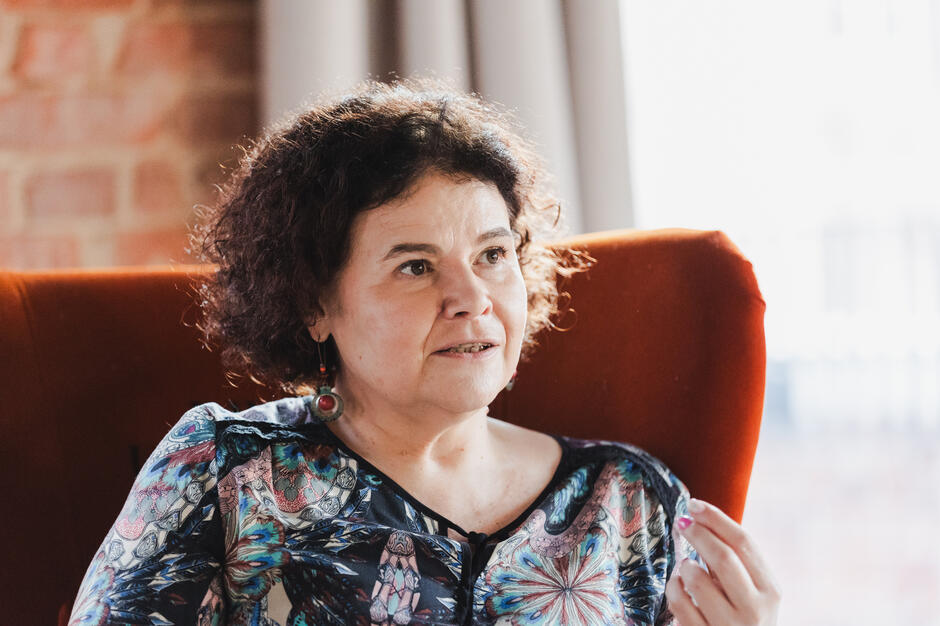 Kobieta z ciemnymi, kręconymi włosami siedzi na pomarańczowym fotelu i prowadzi rozmowę, gestykulując dłonią. Ma na sobie kolorową bluzkę i nosi duże kolczyki, a światło padające z boku tworzy ciepłą, przytulną atmosferę