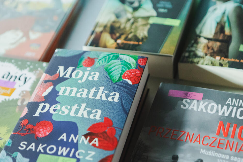 Na stole leży kilka książek, z których najbardziej wyeksponowana to „Moja matka pestka” Anny Sakowicz, z barwną okładką ozdobioną malinami i liśćmi. Obok niej znajdują się inne tytuły autorki, w tym „Nic nie jest przypadkiem”, z widoczną ceną naklejoną na okładce