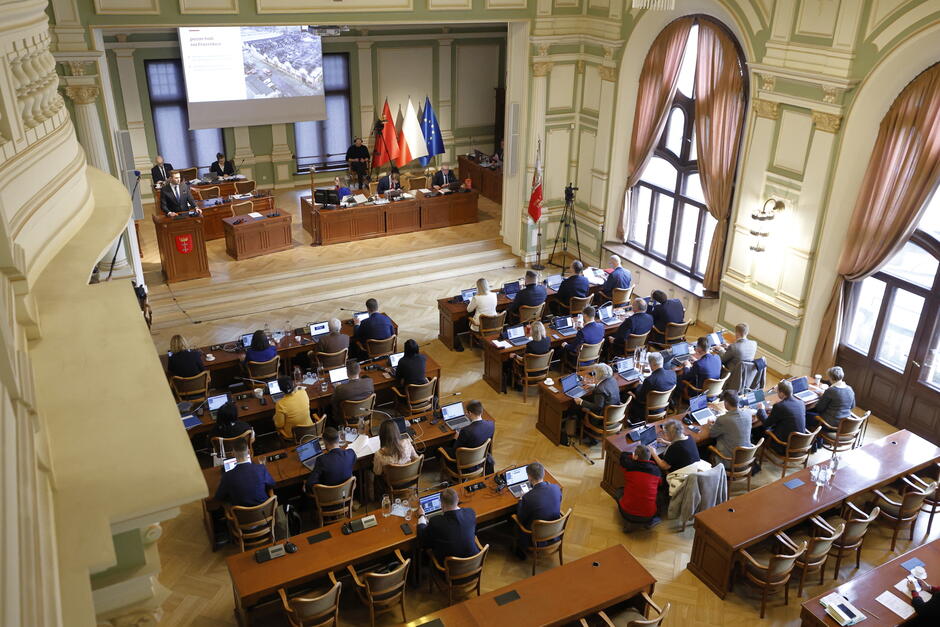 Widok sali obrad z góry, radni siedzą przy stołach ustawionych w długie rzędy