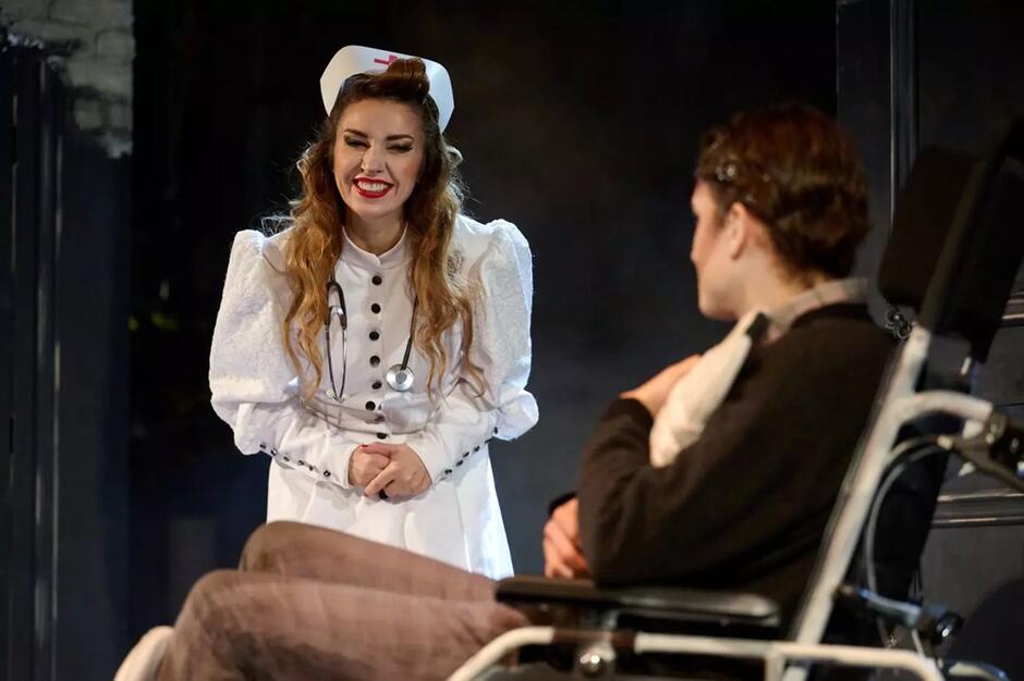 Na zdjęciu widzimy scenę teatralną – kobieta przebrana za uśmiechniętą pielęgniarkę pochyla się w stronę mężczyzny siedzącego na wózku inwalidzkim. Ujęcie pochodzi prawdopodobnie z przedstawienia o nieco groteskowym lub humorystycznym charakterze, co sugeruje przerysowany strój oraz wyrazista mimika aktorki