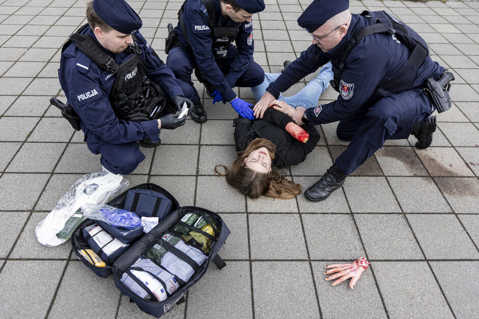 Ćwiczenia w mieście. nad zakrwawioną osobą leżącą na chodniku pochylają się policjanci i opatrują rany. Obok leży sztuczna dłoń.
