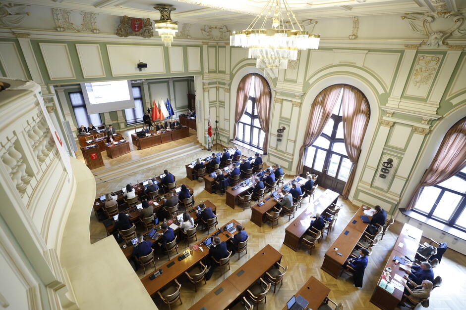 Na zdjęciu widzimy elegancką i reprezentacyjną salę obrad, najprawdopodobniej urzędu miejskiego lub sejmiku wojewódzkiego. Wnętrze jest przestronne, z wysokim sufitem, dużymi, łukowatymi oknami i klasycznymi detalami architektonicznymi w jasnych, pastelowych kolorach. Przez okna wpada dużo naturalnego światła, a całość dopełniają ciężkie, brązowe zasłony. Sala jest wypełniona osobami siedzącymi przy długich stołach – najprawdopodobniej są to radni, urzędnicy oraz zaproszeni goście. Na podwyższeniu znajduje się prezydium z kilkoma osobami prowadzącymi obrady, a za nimi stoją flagi: Polski, Unii Europejskiej oraz najprawdopodobniej lokalne (jedna z nich zawiera czerwone tło i biały gryf, co może wskazywać na Gdańsk).