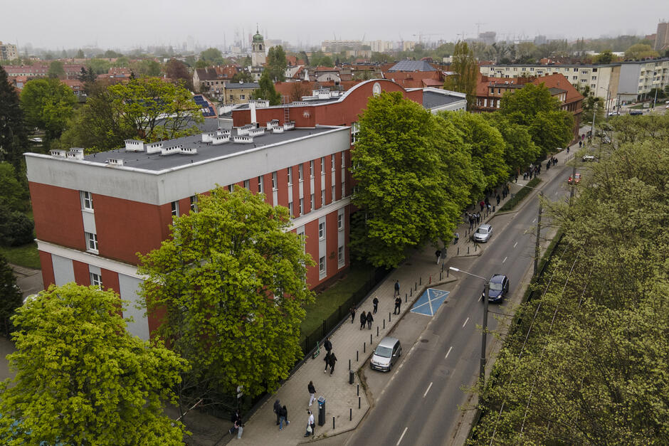 Na zdjęciu widzimy miejską scenę wykonaną z lotu ptaka, prawdopodobnie przy pomocy drona. Centralnym elementem fotografii jest duży budynek o czerwonej i szarej elewacji – wygląda na szkołę lub instytucję edukacyjną. Po lewej stronie kadru znajduje się pas zieleni z dużymi, gęsto rosnącymi drzewami. Drzewa również otaczają budynek od strony chodnika i ulicy. Wzdłuż ulicy po prawej stronie zdjęcia widzimy wiele osób spacerujących, co może sugerować, że to pora wyjścia ze szkoły. Na ulicy poruszają się samochody, a na chodniku widoczna jest niebieska koperta – miejsce parkingowe dla osób z niepełnosprawnością. Pogoda wygląda na pochmurną, a w tle widać zabudowę miejską, z licznymi dachami, wieżami oraz żurawiami portowymi – co może sugerować, że zdjęcie zostało wykonane w Gdańsku.