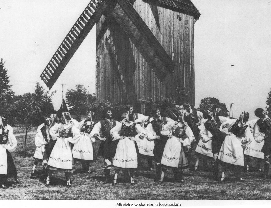 Na zdjęciu widzimy grupę młodzieży ubraną w tradycyjne kaszubskie stroje ludowe. Tancerze – zarówno dziewczęta, jak i chłopcy – ustawieni są w szeregu i wykonują układ taneczny na tle dużego, drewnianego wiatraka. Scena rozgrywa się na otwartej przestrzeni, prawdopodobnie na terenie skansenu, czyli muzeum na wolnym powietrzu. Dziewczęta ubrane są w haftowane fartuszki, długie spódnice, gorsety i charakterystyczne kaszubskie czepki lub chustki. Chłopcy mają na sobie ludowe kamizelki i spodnie, a całość strojów ozdobiona jest typowymi dla Kaszub wzorami haftów – najczęściej w kolorach niebieskim, zielonym, czerwonym i żółtym.  Podpis pod zdjęciem: „Młodzież w skansenie kaszubskim” sugeruje, że fotografia została wykonana podczas wydarzenia promującego kulturę kaszubską – być może podczas święta folkloru, festiwalu lub pokazu dla zwiedzających.