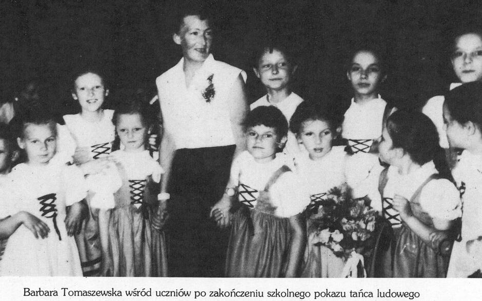 Na zdjęciu widzimy Barbarę Tomaszewską w otoczeniu swoich uczniów, tuż po zakończeniu szkolnego pokazu tańca ludowego. Dzieci ubrane są w tradycyjne stroje ludowe – białe bluzki z bufiastymi rękawami i sznurowane gorsety, które są charakterystycznym elementem wielu polskich regionalnych ubiorów scenicznych. Dziewczynki mają na sobie również długie spódnice, a niektóre z nich trzymają bukiety kwiatów – zapewne jako wyraz podziękowania po występie.