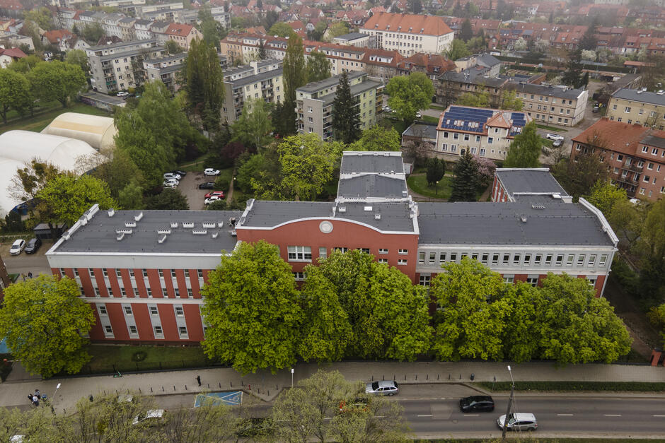 Na zdjęciu widzimy budynek Ogólnokształcącej Szkoły Baletowej im. Janiny Jarzynówny-Sobczak w Gdańsku uchwycony z lotu ptaka. Charakterystyczna bryła szkoły – z czerwonymi i białymi elewacjami oraz półokrągłym zwieńczeniem części środkowej – wyłania się zza gęstej zieleni drzew rosnących od strony ulicy. Przed szkołą znajduje się chodnik i droga z zaparkowanymi samochodami, a w tle – osiedle mieszkaniowe, typowe dla gdańskiej dzielnicy Wrzeszcz. Widoczne po lewej stronie białe namioty to prawdopodobnie obiekty sportowe – być może hale treningowe lub korty. Całość otacza spokojna, miejska zieleń, nadająca temu miejscu kameralny charakter mimo centralnej lokalizacji. To zdjęcie świetnie oddaje atmosferę szkoły – solidnej, zakorzenionej w historii, ale otoczonej energią i ruchem miasta. Chcesz, żebym przygotował podpis albo opis do folderu promocyjnego, jubileuszu lub wystawy?