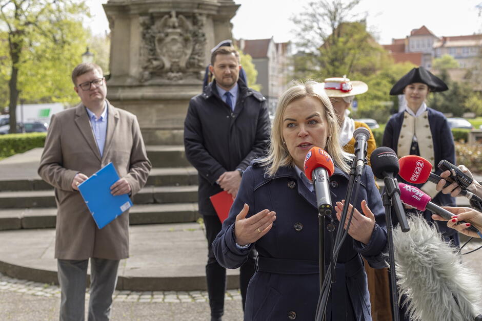 Kobieta około lat 35, o blond włosach, przemawia do mikrofonów