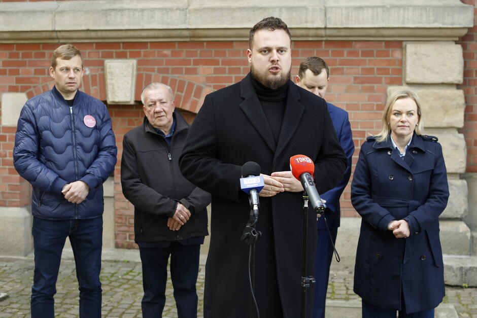  Grupa pięciu osób stojących przed ceglanym budynkiem. Osobą w środku jest mężczyzna z brodą i mówi do wielu mikrofonów. Ma na sobie czarny płaszcz i wydaje się, że zwraca się do mediów. Za nim są cztery inne osoby, dwóch mężczyzn i dwójka kobiet, które również uważnie słuchają mówiącego męża. Po lewej stronie stoi z rękami przymocowanymi do siebie, podczas gdy pozostałe dwie stoją za nim. Wszyscy patrzą na kamerę i wyglądają poważnie.
