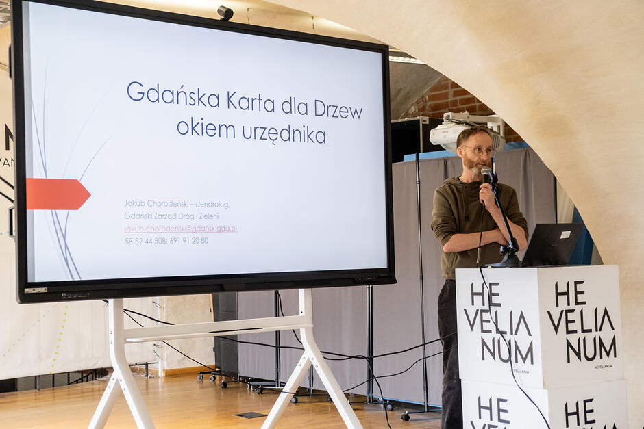 W sali konferencyjnej w starym budynku z łukowym sklepieniem stoi przy mównicy mężczyzna i mówi do mikrofonu. Obok stoi ekran z prezentacją.