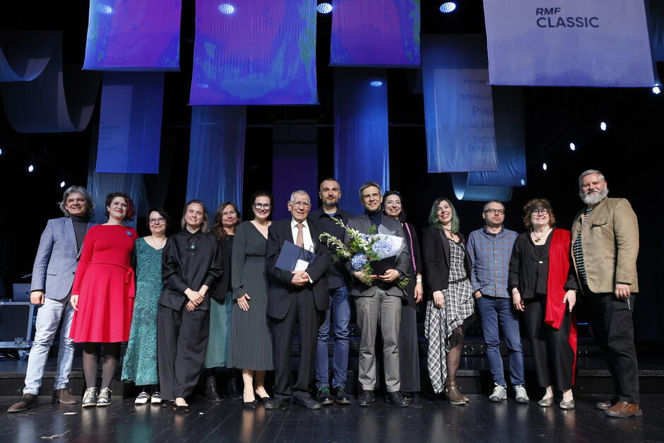 Cała grupa laureatów, organizatorów i gości pozuje wspólnie na scenie, celebrując uroczystość. Na twarzach uczestników widać radość i dumę, a tło sceny zdobią podświetlone transparenty z nazwami patronów