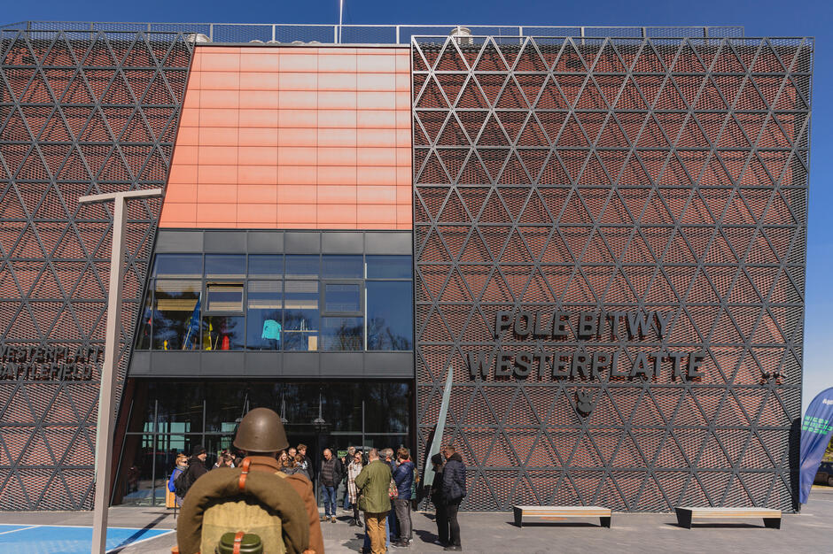 Na zdjęciu widać nowoczesny budynek muzeum „Pole Bitwy Westerplatte” w Gdańsku, z charakterystyczną fasadą wykonaną z czerwonych paneli i metalowych trójkątnych konstrukcji. Przed wejściem zgromadzili się zwiedzający, w tym osoba w historycznym mundurze wojskowym.
