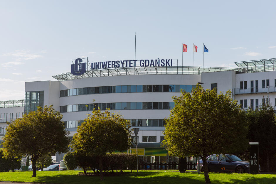 Duży biały budynek z niebieskim znakiem, na którym jest napisane "UNIVERSYTET GDANSKI". Z przodu znajdują się trzy drzewa, a obok drzew na trawie zaparkowany jest czarny samochód. Za płotem znajduje się czarny płot. Dwie flagi są położone jedna na drugiej.