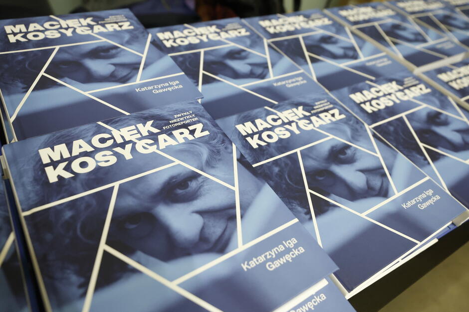 Na zdjęciu widać stos egzemplarzy książki zatytułowanej „Maciek Kosycarz. Zwykły Niezwykły Fotoreporter”, autorstwa Katarzyny Igi Gawęckiej. Okładki książek są w odcieniach niebieskiego i przedstawiają portret fotograficzny Macieja Kosycarza podzielony białymi liniami na geometryczne kształty. 