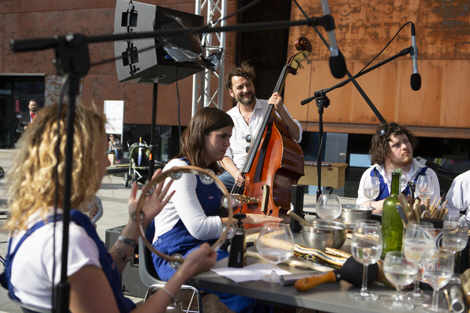 Grupa muzyków występuje na świeżym powietrzu, używając nietypowych instrumentów, takich jak naczynia kuchenne i kieliszki, a w tle widać nowoczesną, rdzawą architekturę. Jeden z muzyków gra na kontrabasie, a inni koncentrują się na grze na instrumentach perkusyjnych i tamburynie.