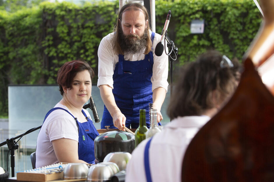 W kameralnym ustawieniu na świeżym powietrzu, muzyk z długą brodą gra na metalowych miskach, podczas gdy młoda kobieta w niebieskich ogrodniczkach spogląda w stronę kamery z lekkim uśmiechem. W tle widoczna jest zielona ściana roślinności, tworząc przyjazną atmosferę występu