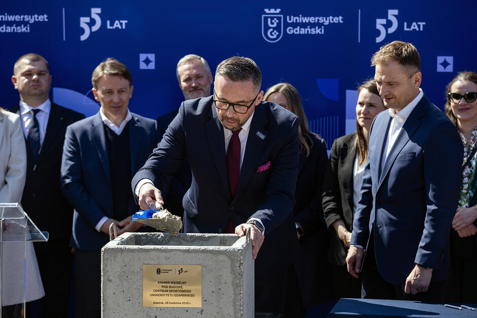 Dwóch mężczyzn stoi przy miejscu wmurowania kamienia węgielnego pod budowę. Jeden z nich wrzuca beton na kamień węgielny. Za nimi stoi grupa innych osób.