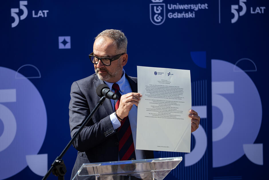 Mężczyzna w garniturze stoi przy mównicy i pokazuje akt erekcyjny podczas uroczystości wmurowania kamienia węgielnego na budowie.