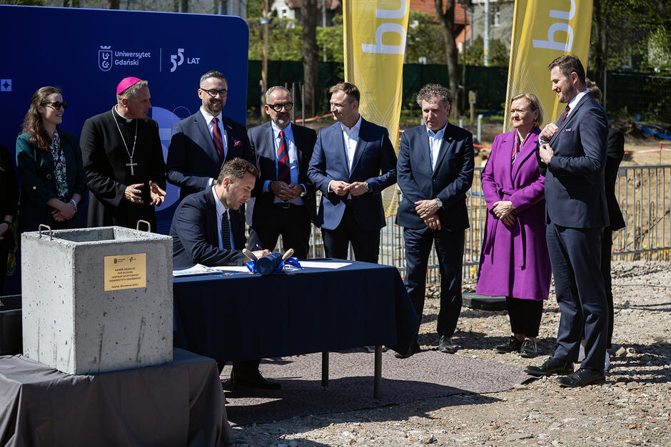 Mężczyzna siedzi przy stole i podpisuje akt erekcyjny budowy Centrum Sportowego, obok niego stoi kilka innnych osób biorących udział w wydarzeniu.