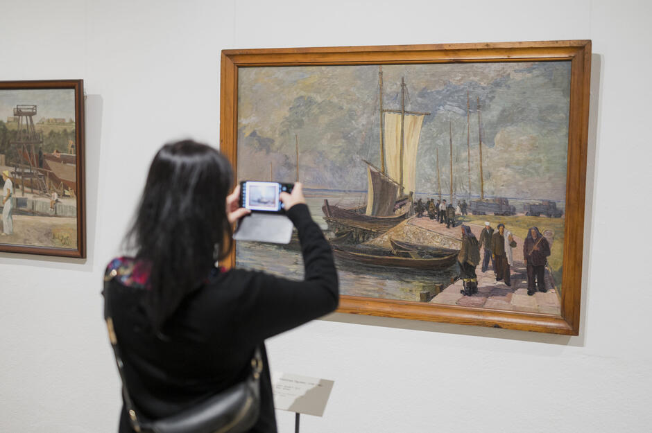 Zdjęcie przedstawia osobę fotografującą obraz wiszący w galerii sztuki – dzieło ukazuje port z żaglówkami, tłumem ludzi na nabrzeżu i malowniczym niebem w tle. W tle widoczny jest także drugi obraz o tematyce industrialnej, co wskazuje na ekspozycję związaną z tematyką pracy lub życia codziennego w epoce socrealizmu