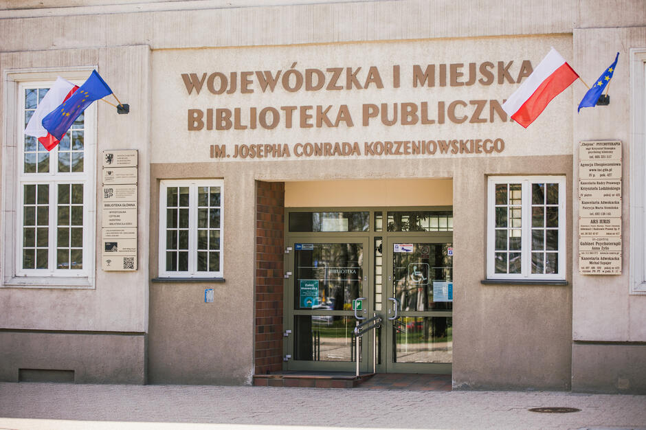 Zdjęcie [rzedstawia wejście do Wojewódzkiej i Miejskiej Biblioteki Publicznej im. Josepha Conrada-Korzeniowskiego. Na budynku widoczne są flagi Polski i Unii Europejskiej oraz tablice informacyjne