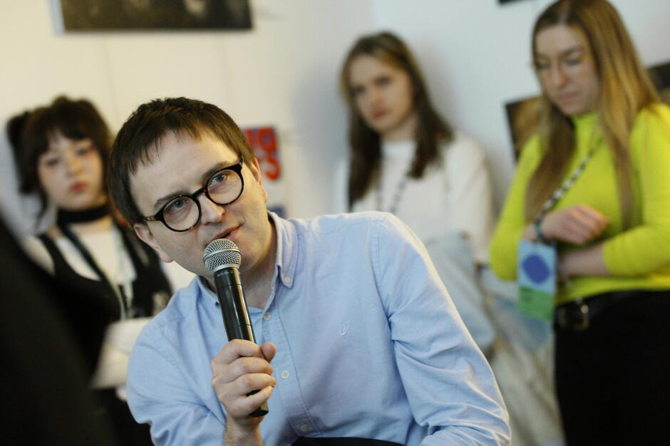 Mężczyzna w jasnoniebieskiej koszuli i okrągłych okularach mówi do mikrofonu podczas wydarzenia kulturalnego. W tle widać kilka słuchających kobiet