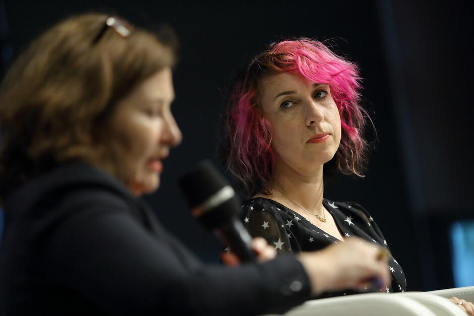 Kobieta z różowo-czarnymi włosami i sukienką w gwiazdki patrzy z zainteresowaniem na mówiącą osobę z mikrofonem. Scena ma charakter panelu dyskusyjnego