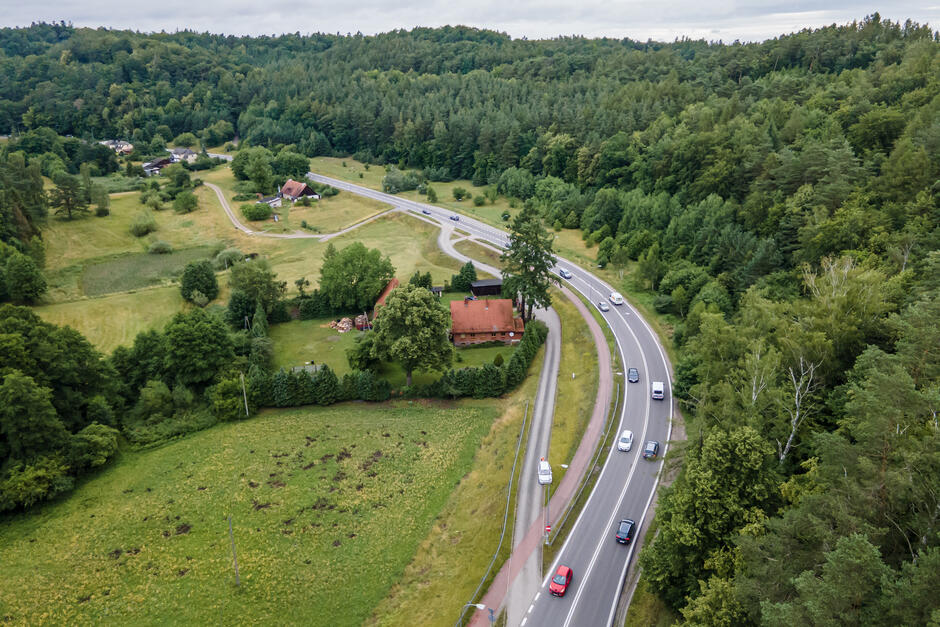 Zdjęcie z lotu ptaka. Wśród porośniętych lasem wzgórz morenowych wije się ulica, na której jada samochody. Obok ulicy stoi dom z dachem krytym dachówką.