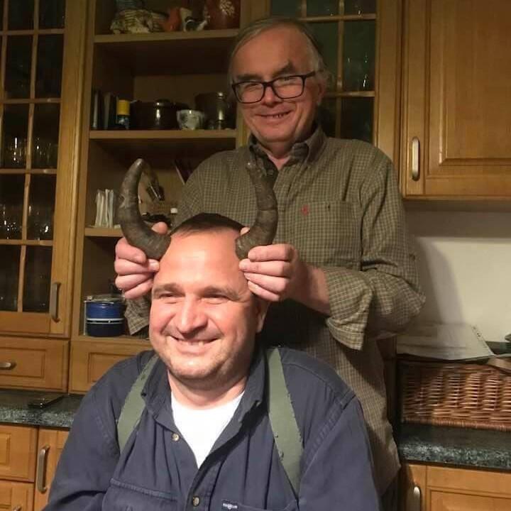 Dwaj mężczyźni żartują sobie. Jeden siedzi na krześle, zwrócony twarzą do aparatu fotograficznego, drugi stoi za nim - przykłada temu pierwszemu bycze rogi, po obu stronach głowy