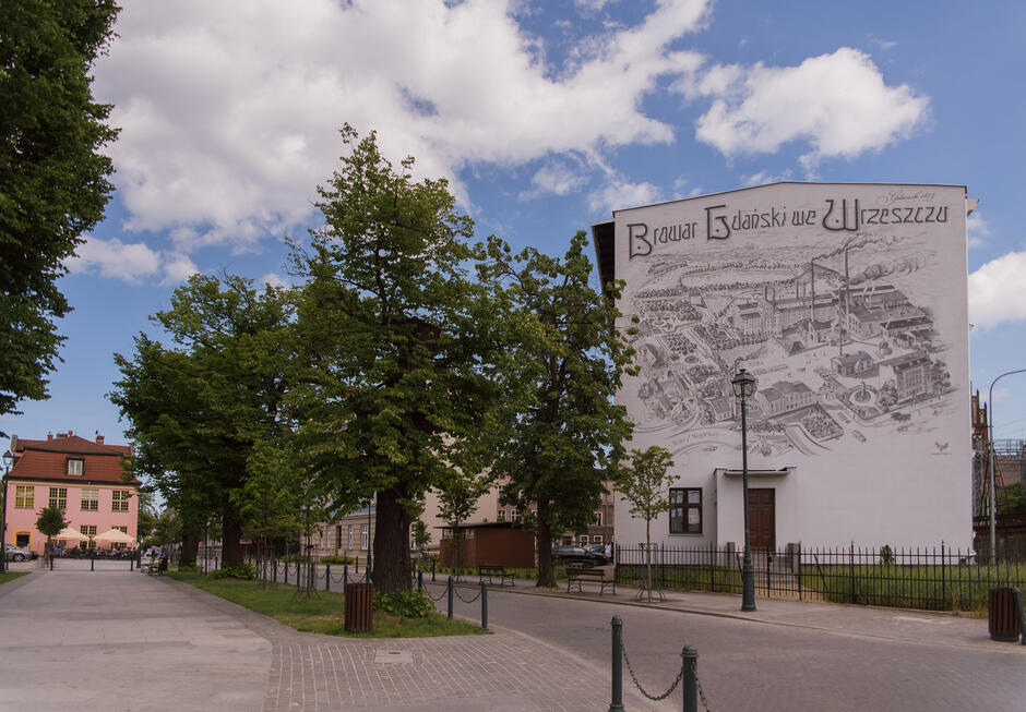 Na boku stoi duży biały znak z czarnym ogrodzeniem za znakiem. Na znaku jest czarno-biały rysunek i napisane na nim białymi literami słowo BEWAR . Czarne światło uliczne znajduje się na szarym słupie obok ogrodzenia. Poniżej chodnika widać część utwardzonego chodnika. Wysokie zielone drzewa stoją w trawie w pobliżu chodnika, na którym są szare metalowe słupy z łańcuchami.