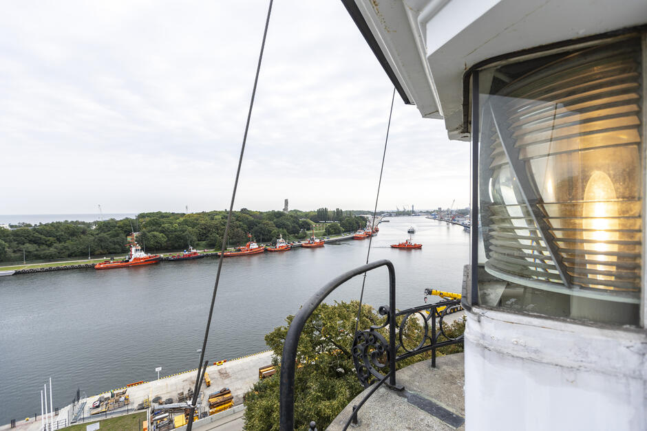 widok z szczytu latarni, patrząc w dół na port; port jest wypełniony kilkoma czerwonymi i pomarańczowymi łodziami, a w wodzie jest kilka żurawi i innych statków; po prawej stronie biała latarnia z szklanym oknem i metalową szyną; w tle widzimy panoramę miasta z budynkami i drzewami na brzegu; nad portem niebo jest przeludnione.