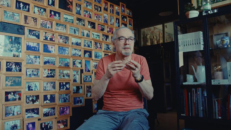  Starszy mężczyzna w okularach i czerwonej koszulce siedzi przed ścianą pokrytą dziesiątkami oprawionych zdjęć. Wygląda na zamyślonego lub zaangażowanego w rozmowę, gestykulując dłońmi