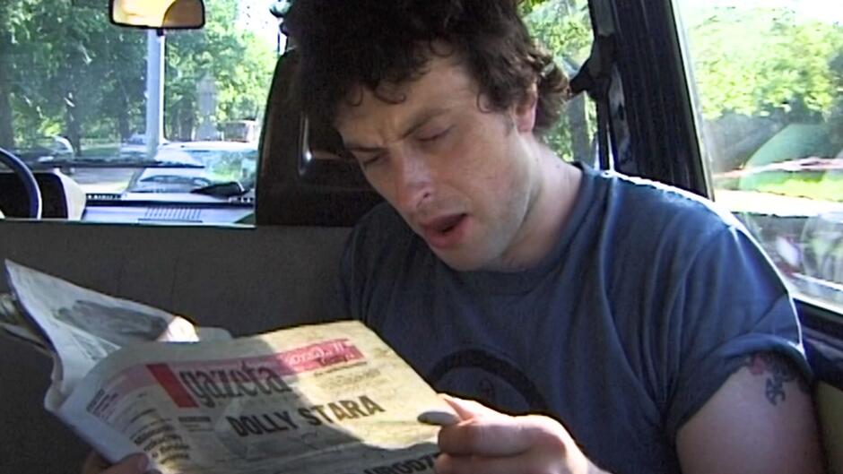 Młody mężczyzna siedzi w samochodzie i z zaangażowaniem czyta gazetę. Jego wyraz twarzy wskazuje na zaskoczenie lub skupienie.