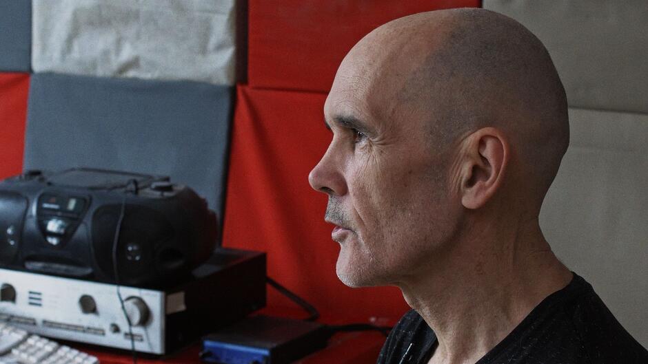 Profil łysego mężczyzny, który siedzi w pomieszczeniu z kolorowymi panelami akustycznymi na ścianie. Obok niego znajduje się stara wieża stereo i klawiatura, co sugeruje otoczenie studyjne lub muzyczne