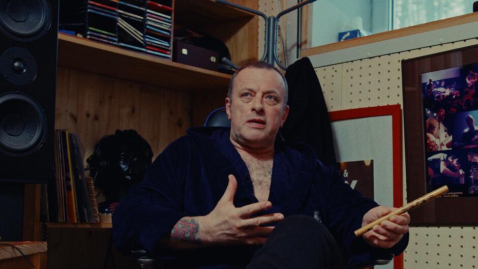 Mężczyzna z tatuażem na przedramieniu siedzi w studiu muzycznym trzymając pałeczkę perkusyjną. Jego poza i gestykulacja sugerują, że opowiada z pasją o czymś ważnym.
