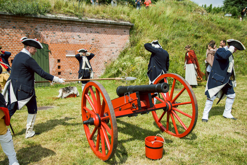 Na zdjęciu przedstawiona jest inscenizacja historyczna z udziałem rekonstruktorów ubranych w XVIII-wieczne mundury, przygotowujących się do wystrzału z czerwono-czarnej armaty. Część uczestników zakrywa uszy, co sugeruje, że moment odpalenia działa jest bliski.