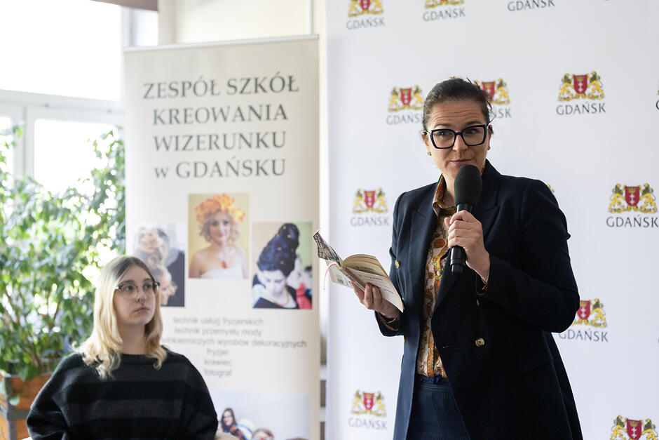 Kobieta stoi w sali i trzymając książkę mówi do zebranych, których na zdjęciu nie widać. Obok siedzi jedna uczennica.