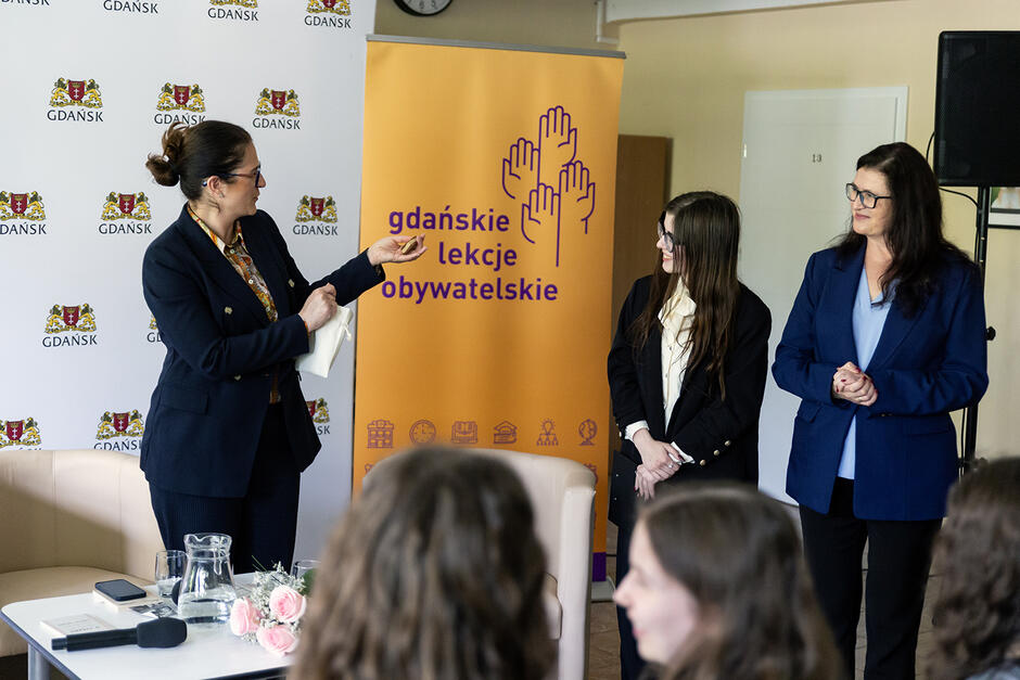 W sali stoja dwie dorosłe kobiet i jedna uczennica i rozmawiają. Przed nimi siedzą na krzesłach uczniowie.