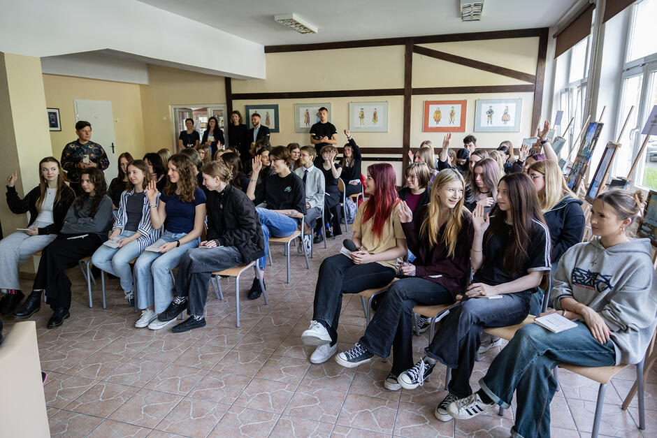 W dużej sali siedzą na krzesłach uczniowie, głównie dziewczyny. Niektóre śmieją się.