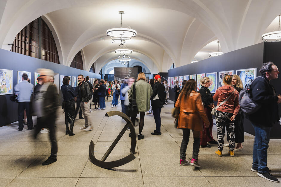 W przestronnej galerii z łukowatym sufitem tłum ludzi ogląda kolorowe obrazy rozmieszczone po obu stronach korytarza. W centralnej części widać abstrakcyjną rzeźbę w kształcie przeciętego okręgu, a odwiedzający żywo rozmawiają i podziwiają dzieła.